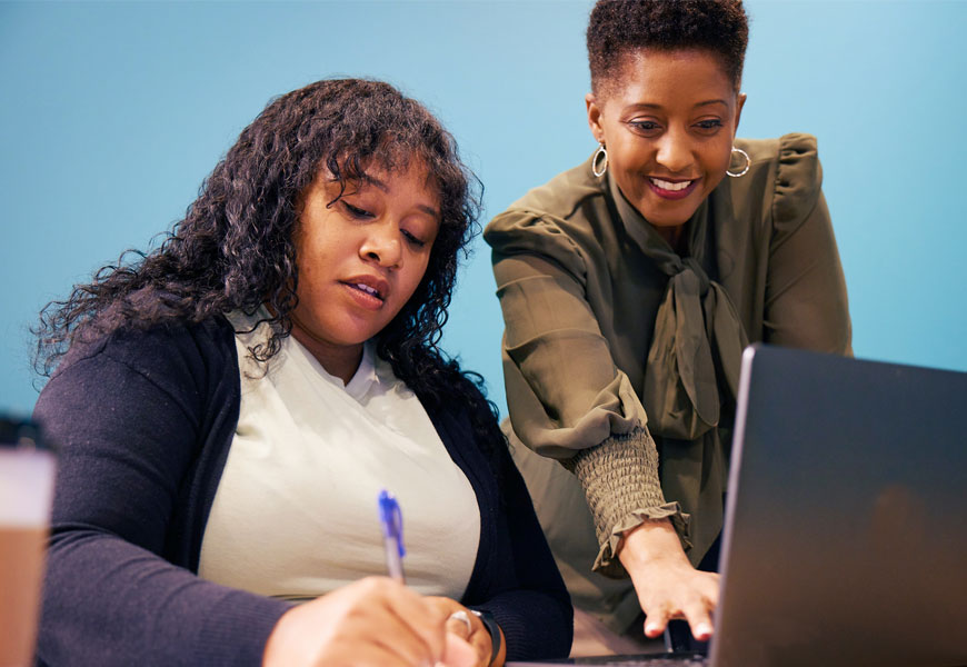 Woman smiling facing a laptop while another seated woman takes notes. 
