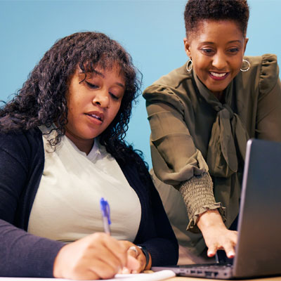 Woman smiling facing a laptop while another seated woman takes notes. 