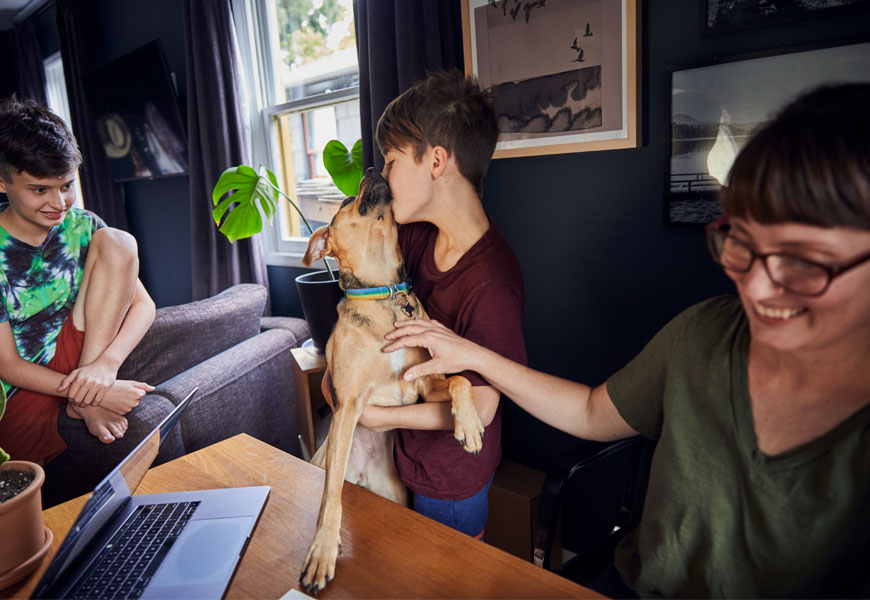 Two children playing with a dog in front of a desk with a laptop. 